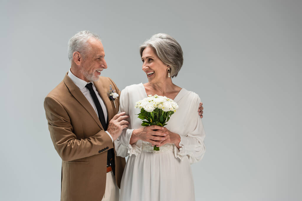 happy mature bride in wedding dress holding bouquet and standing with middle aged groom in suit isolated on grey - Foto, Imagen