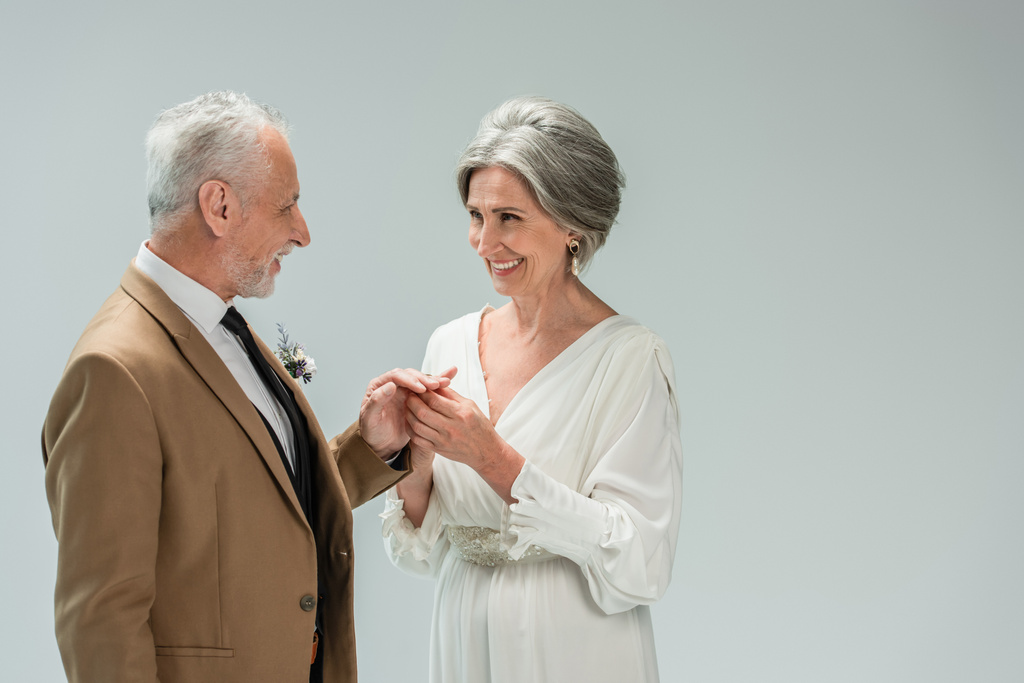 happy mature bride in white dress wearing engagement ring on finger of middle aged groom isolated on grey - Foto, Imagem