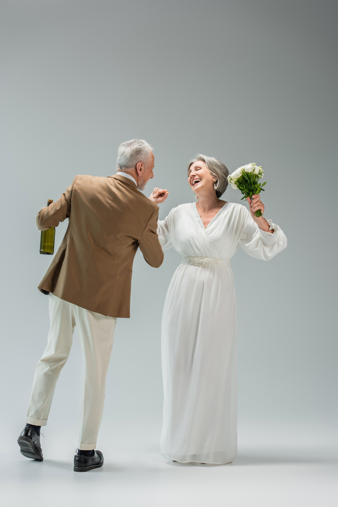 full length of happy middle aged man holding bottle of champagne while dancing with cheerful bride in white dress on grey - Foto, Bild