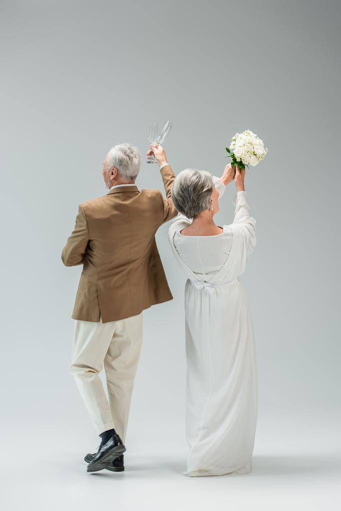 back view of middle aged man holding champagne glasses near bride in white dress with wedding bouquet on grey - Zdjęcie, obraz
