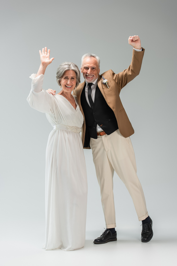 full length of happy mature newlyweds waving hands standing smiling and standing on grey  - Foto, Bild