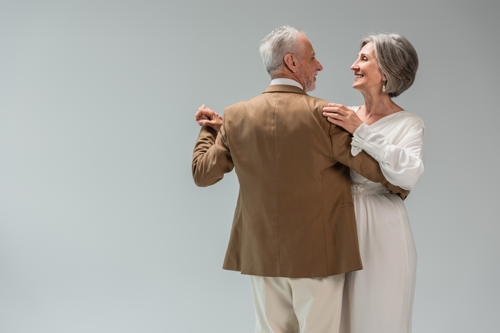 Ευτυχισμένος μεσήλικας γαμπρός και νύφη με νυφικό να κρατιούνται χέρι χέρι ενώ χορεύουν απομονωμένοι σε γκρι  - Φωτογραφία, εικόνα