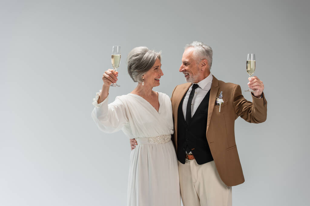 cheerful middle aged groom and happy bride in white dress holding glasses of champagne isolated on grey - Foto, afbeelding