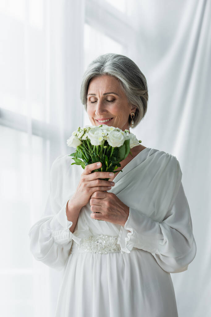 happy middle aged bride in white dress smelling wedding bouquet near curtains  - Valokuva, kuva