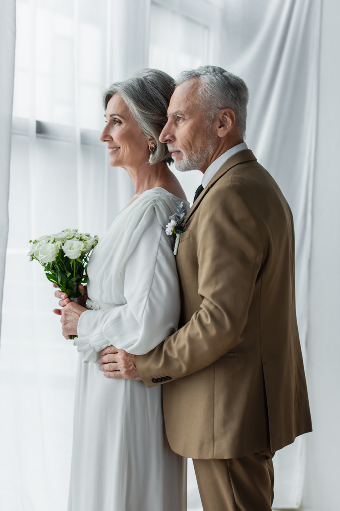 бородатий середнього віку наречений обіймає щасливу наречену в білій сукні з весільним букетом
  - Фото, зображення