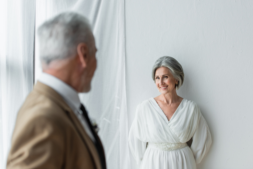 blurred man in suit looking at cheerful mature bride in white dress near white curtains - Fotografie, Obrázek