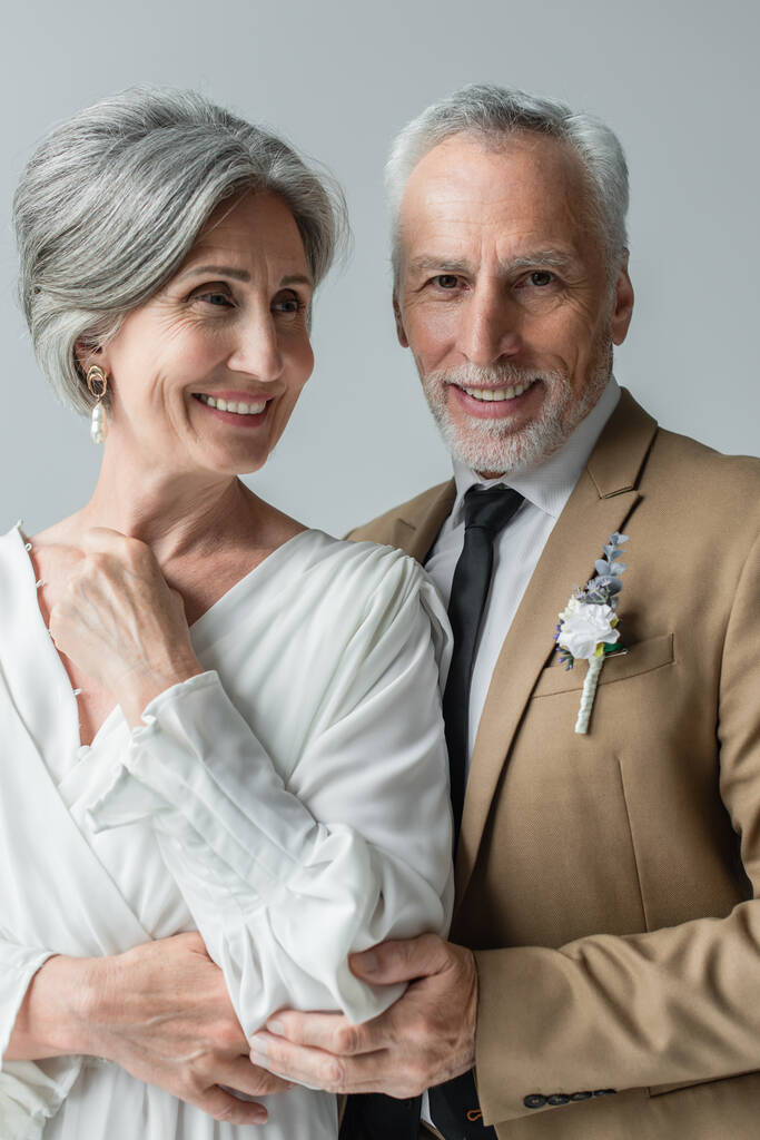 happy middle aged man in suit with floral boutonniere and woman in white wedding dress isolated on grey - Zdjęcie, obraz