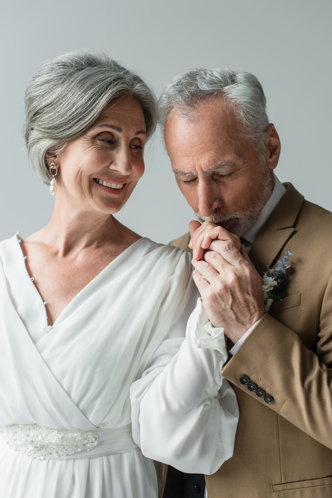 middle aged man in suit kissing hand of happy bride in white wedding dress isolated on grey - Photo, Image