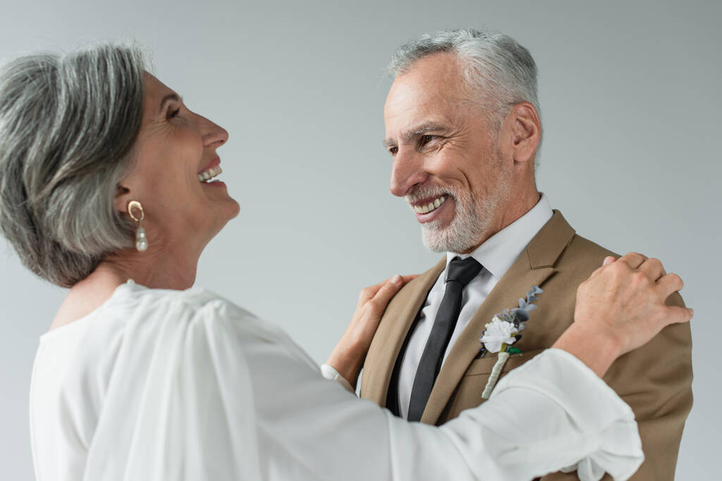 happy middle aged bride in white wedding dress hugging bearded groom and laughing isolated on grey - Fotó, kép