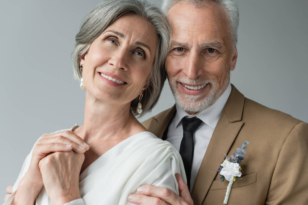 portrait of happy middle aged man in suit with floral boutonniere hugging woman in white wedding dress isolated on grey - Fotoğraf, Görsel