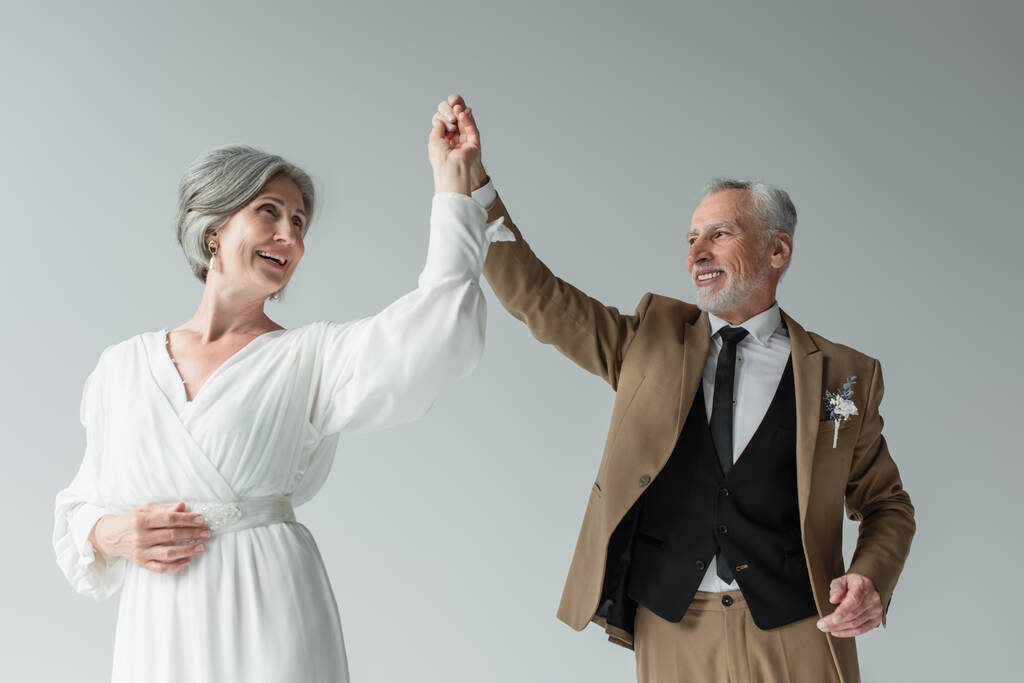 щасливий чоловік середнього віку в костюмі, танцює зі усміхненою нареченою в білій весільній сукні ізольовано на сірому
 - Фото, зображення