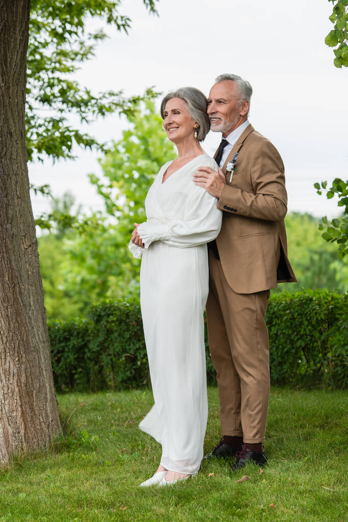 full length of happy middle aged groom in suit hugging mature bride in white dress in garden  - Photo, Image
