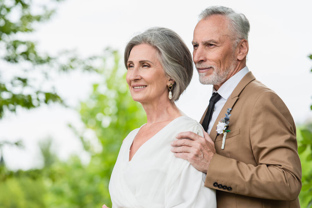 задоволений наречений середнього віку в костюмі обіймає зрілу наречену в білій сукні в саду
  - Фото, зображення