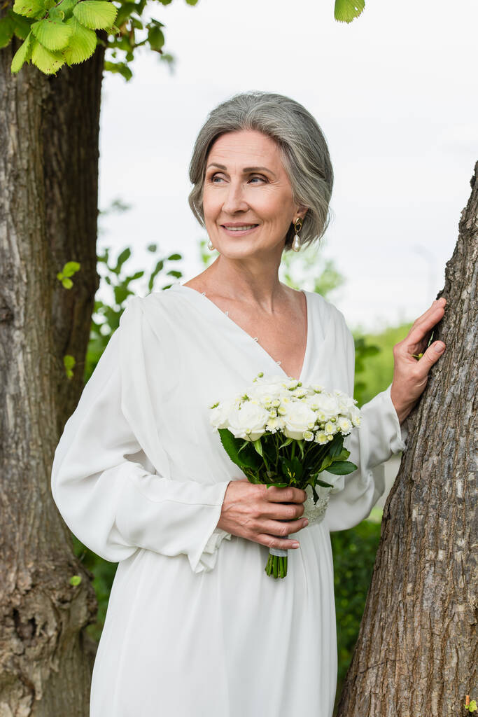 pleased middle aged bride in white dress holding wedding bouquet near tree trunk in park  - Foto, afbeelding