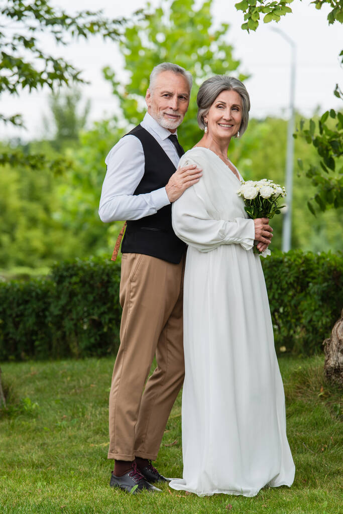 full length of pleased mature man in formal wear hugging bride with wedding bouquet in garden - 写真・画像