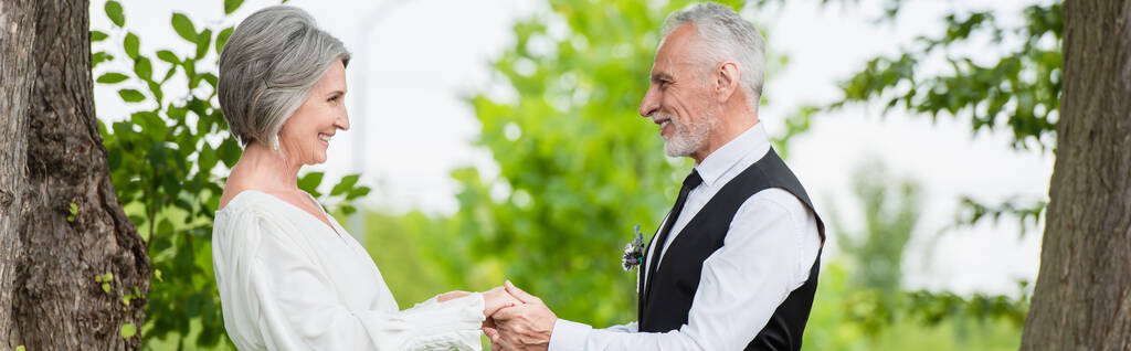 вид збоку веселий зрілий чоловік у формальному одязі тримає руки з усміхненою нареченою у весільній сукні в саду, банер
 - Фото, зображення
