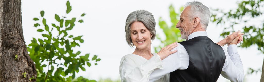 щасливий чоловік середнього віку в офіційному одязі танцює з нареченою в білій весільній сукні в зеленому саду, банер
 - Фото, зображення