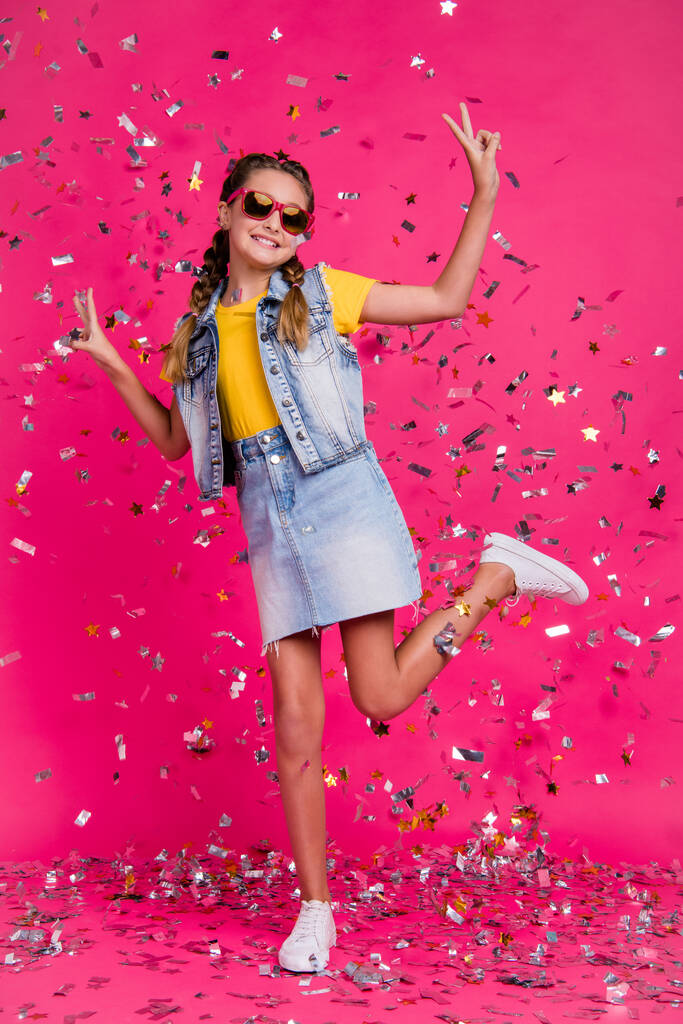 Полноразмерная вертикальная фотография маленькой девочки с V-образным знаком, в очках, футболке и юбках, изолированных на ярком цветовом фоне. - Фото, изображение