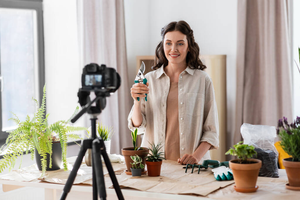 bloggen, tuinieren en huishoudelijk werk concept - gelukkige vrouw of blogger met camera en snoeier planten pot bloemen en opnemen tutorial video thuis - Foto, afbeelding