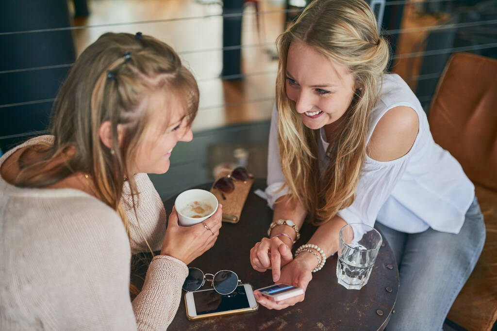 Catching up over coffee. High angle shot of two attractive young girlfriends looking at a cellphone while sitting in a cafe - Fotó, kép