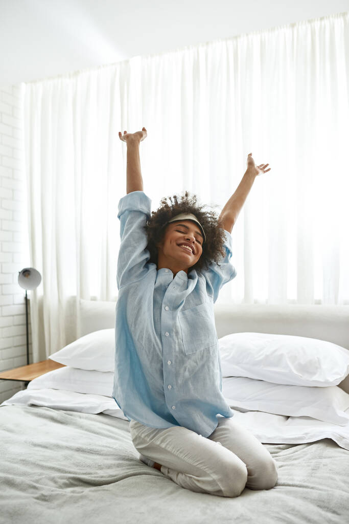 Young smiling black girl waking up on bed at home in morning time. Curly teen female zoomer with closed eyes wearing homewear and sleep mask. Domestic lifestyle. Modern bedroom in spacious apartment - Fotografie, Obrázek