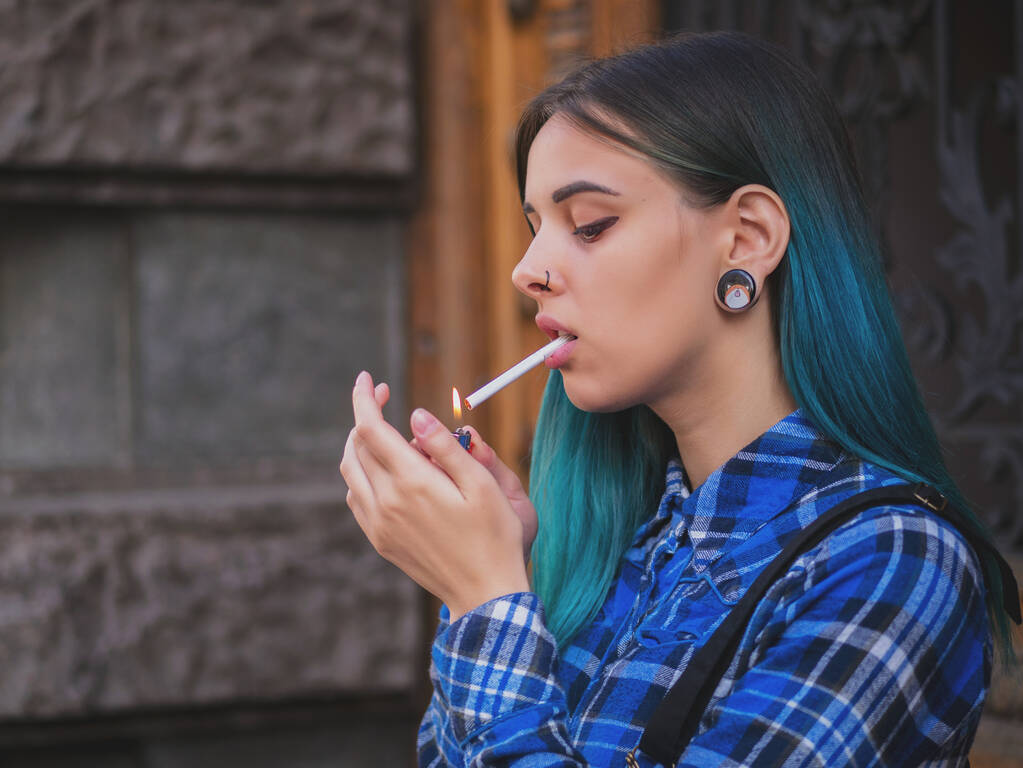 Chica punk joven fumando cigarrillo junto a la puerta de madera.Hipster con el pelo teñido de azul. Mujer con piercing en nariz, lentes violetas, orejas túneles y peinado inusual
. - Foto, imagen