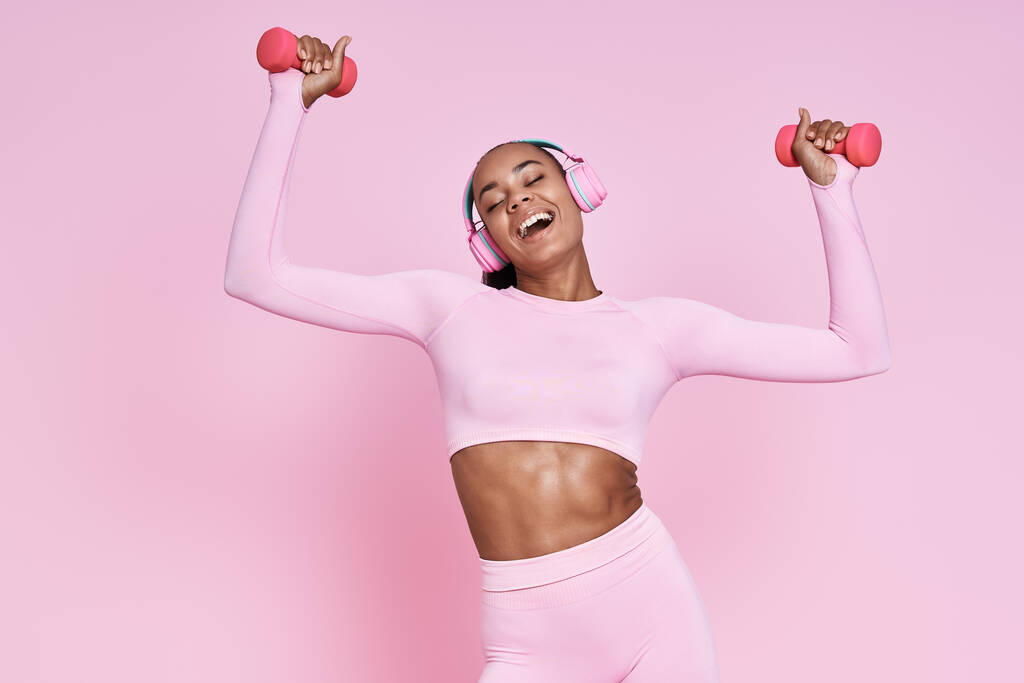 Beautiful woman in headphones exercising with dumbbells and smiling against pink background - Fotó, kép