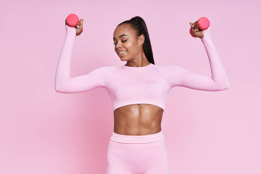 Beautiful woman exercising with dumbbells and smiling while standing against pink background - Valokuva, kuva