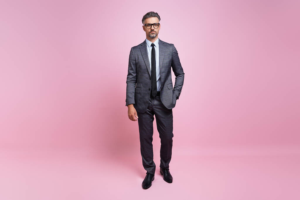 Confident mature man in full suit looking at camera while standing against pink background - 写真・画像