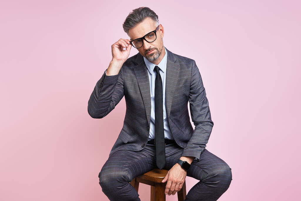 Handsome mature man in formalwear adjusting eyeglasses while sitting against pink background - Foto, Imagen