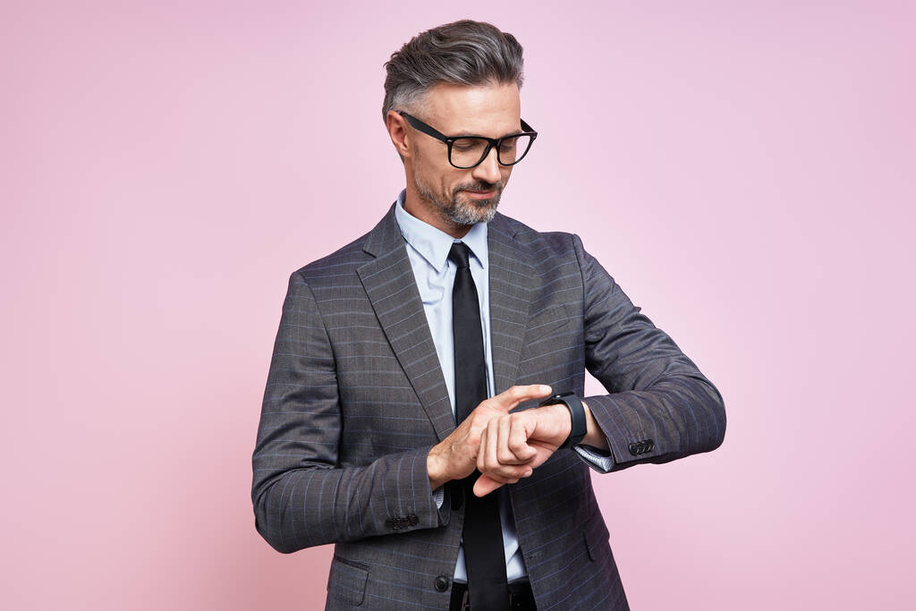 Confident mature man in formalwear checking the time while standing against pink background - Foto, immagini