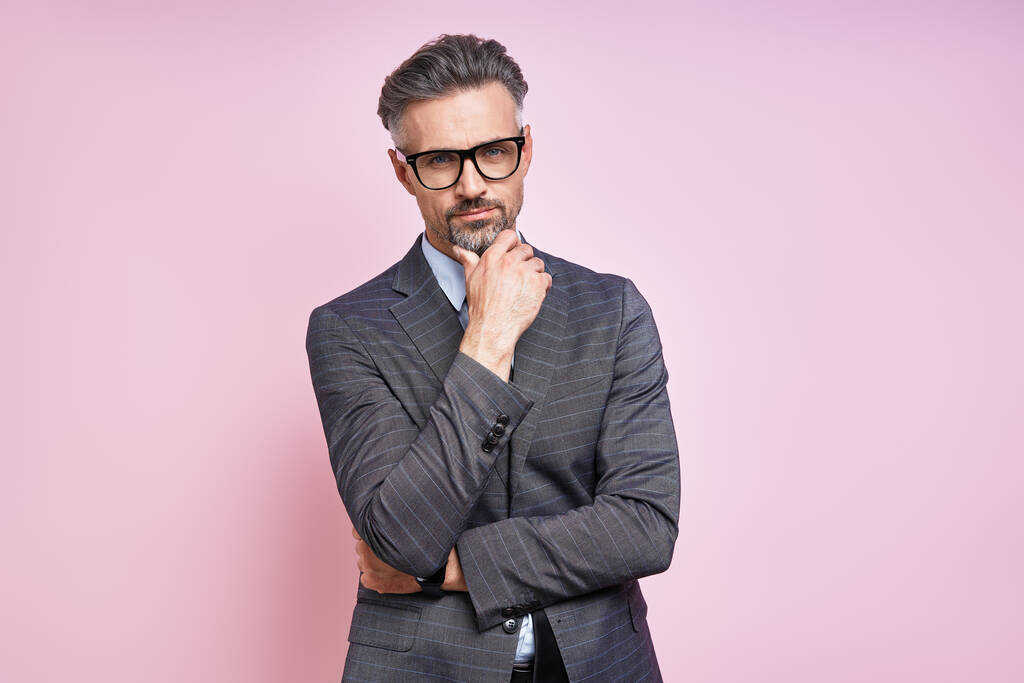 Handsome mature man in formalwear holding hand on chin while standing against pink background - Foto, afbeelding