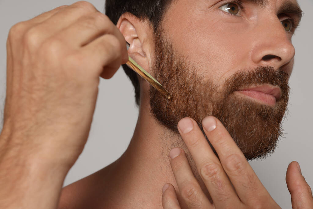 Man applying oil onto beard on grey background, closeup - Fotografie, Obrázek