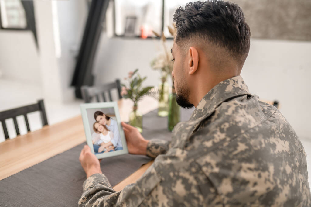 Man in the camouflage uniform sitting at the kitchen table and holding the photo in his hands - Fotoğraf, Görsel
