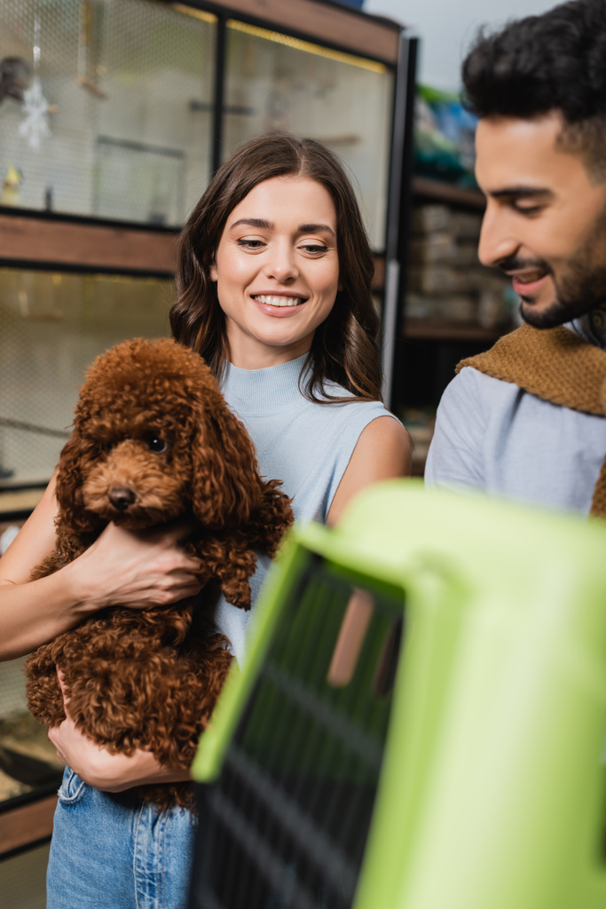 Smiling woman holding poodle near arabian boyfriend and cage in animal shop - Photo, Image