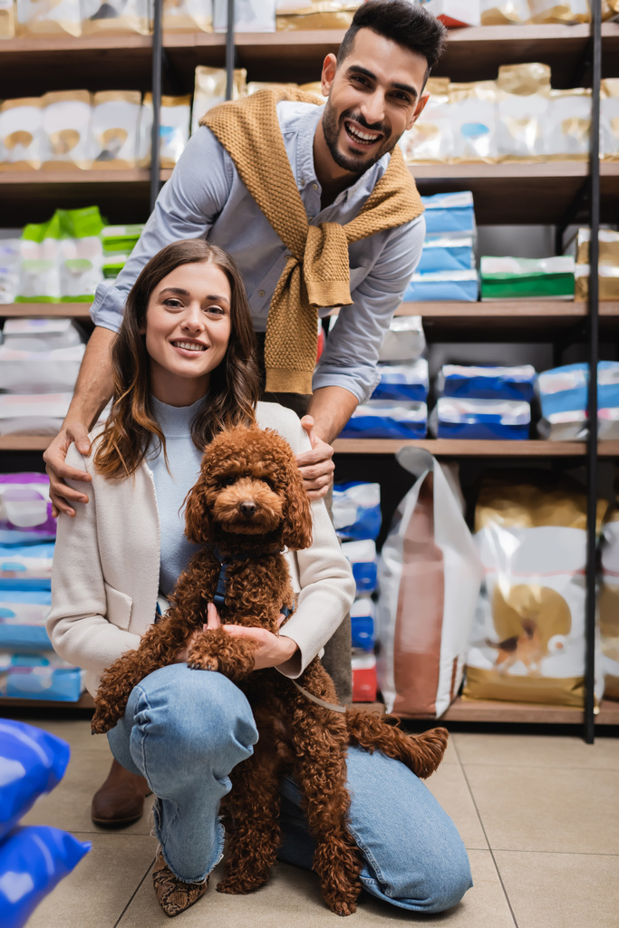 Cheerful interracial couple with brown poodle looking at camera in pet shop  - Fotó, kép