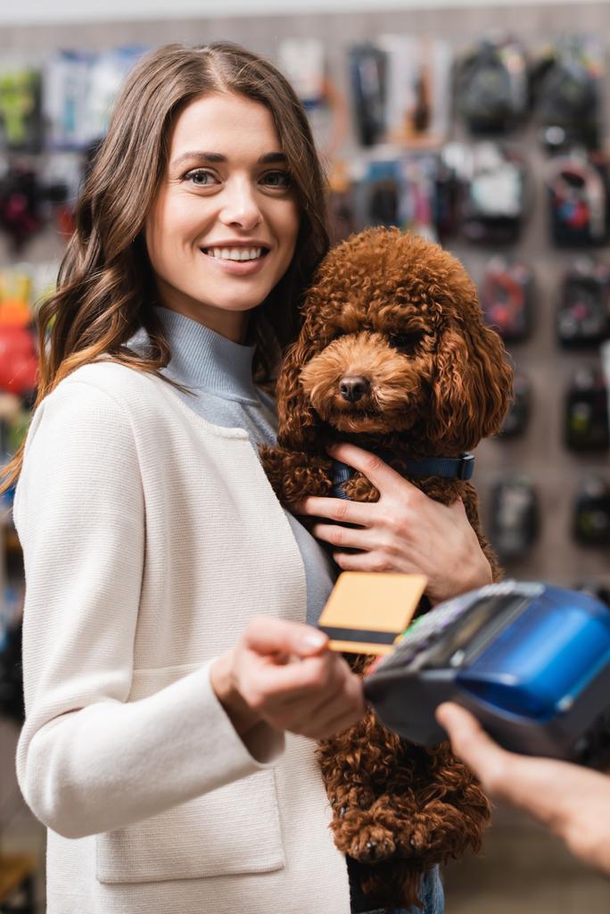 Cheerful woman holding poodle and credit card near payment terminal in pet shop - Foto, afbeelding