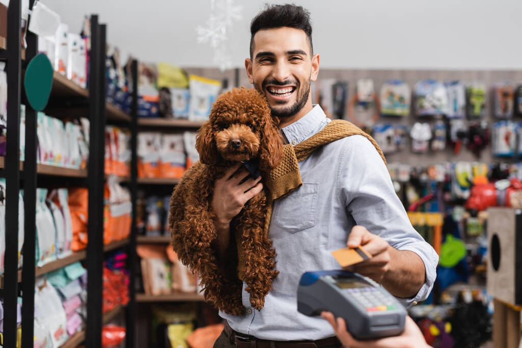 Positive arabian man holding poodle and looking at camera while paying with credit card in pet shop  - 写真・画像