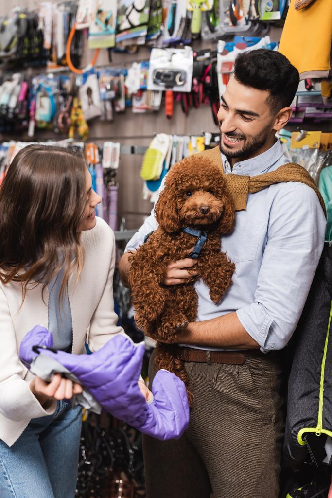 Cheerful interracial couple with poodle choosing animal jacket in pet shop  - Foto, immagini