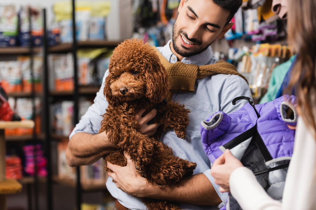 Blurred woman holding animal jacket near arabian boyfriend with brown poodle in pet shop  - Foto, afbeelding