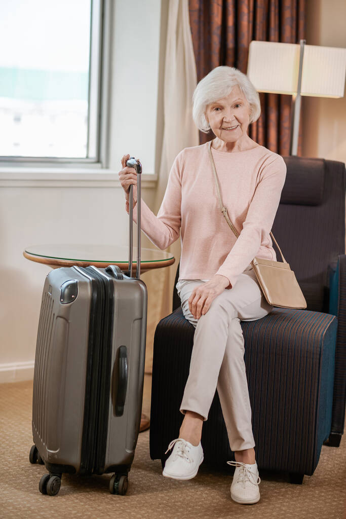 In a hotel. Good-looking senior woman sitting with a suitcase in a hotel room - Foto, Imagen