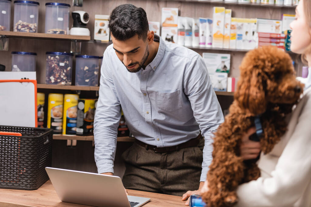 Arabian salesman looking at laptop near blurred customer with poodle in pet shop  - Fotó, kép