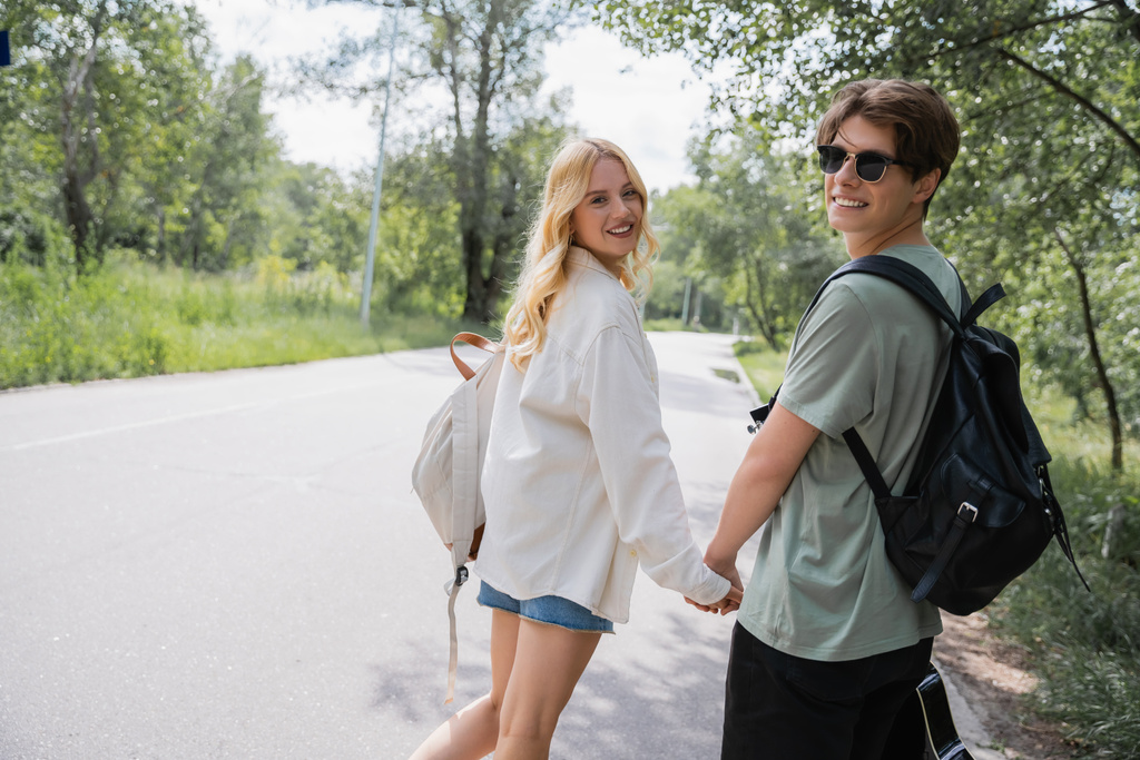 ευτυχισμένο νεαρό ζευγάρι με σακίδια που κρατιούνται χέρι-χέρι και κοιτάζουν την κάμερα στον επαρχιακό δρόμο - Φωτογραφία, εικόνα