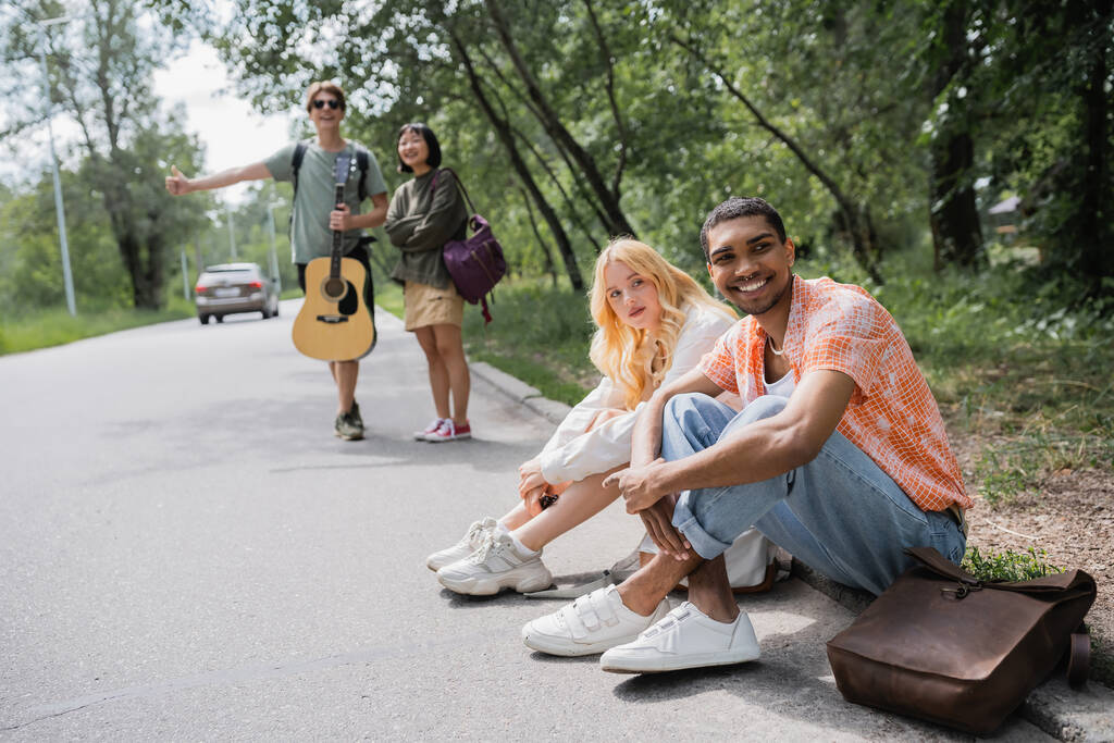 blurred hitchhikers stopping car near smiling interracial friends sitting on road - Foto, imagen
