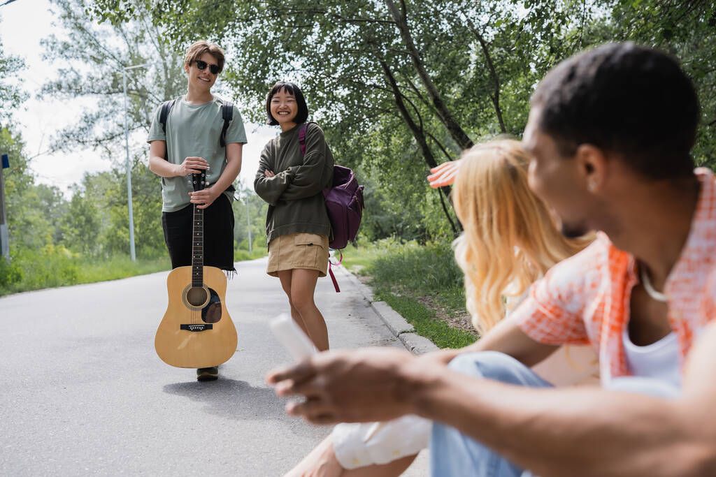усміхнений чоловік тримає гітару біля азіатської жінки і розмиті міжрасові друзі сидять на дорозі
 - Фото, зображення