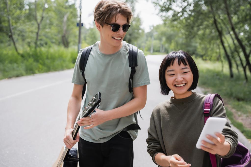 smiling asian woman searching direction on smartphone near young man in sunglasses - Photo, image