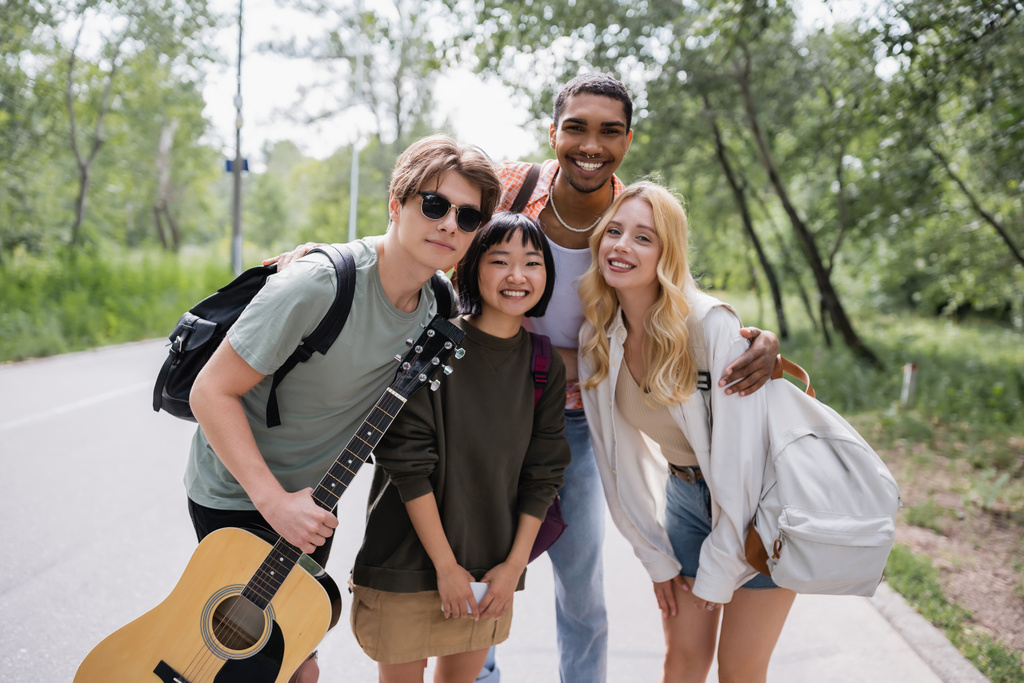 joyful multicultural friends with backpacks and guitar smiling at camera on countryside road - Fotoğraf, Görsel