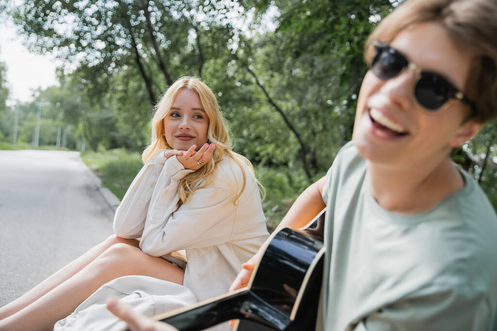 blonde woman looking at happy man playing guitar on blurred foreground - Foto, Imagen