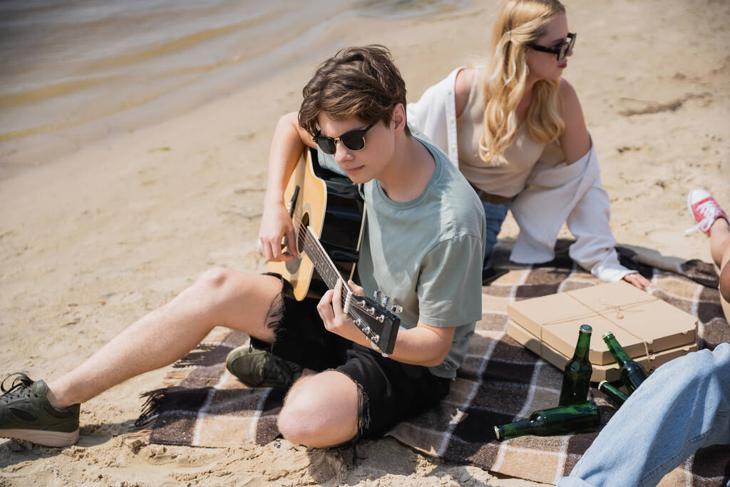 άνδρας με γυαλιά ηλίου που παίζει ακουστική κιθάρα κατά τη διάρκεια πάρτι στην παραλία με φίλους - Φωτογραφία, εικόνα