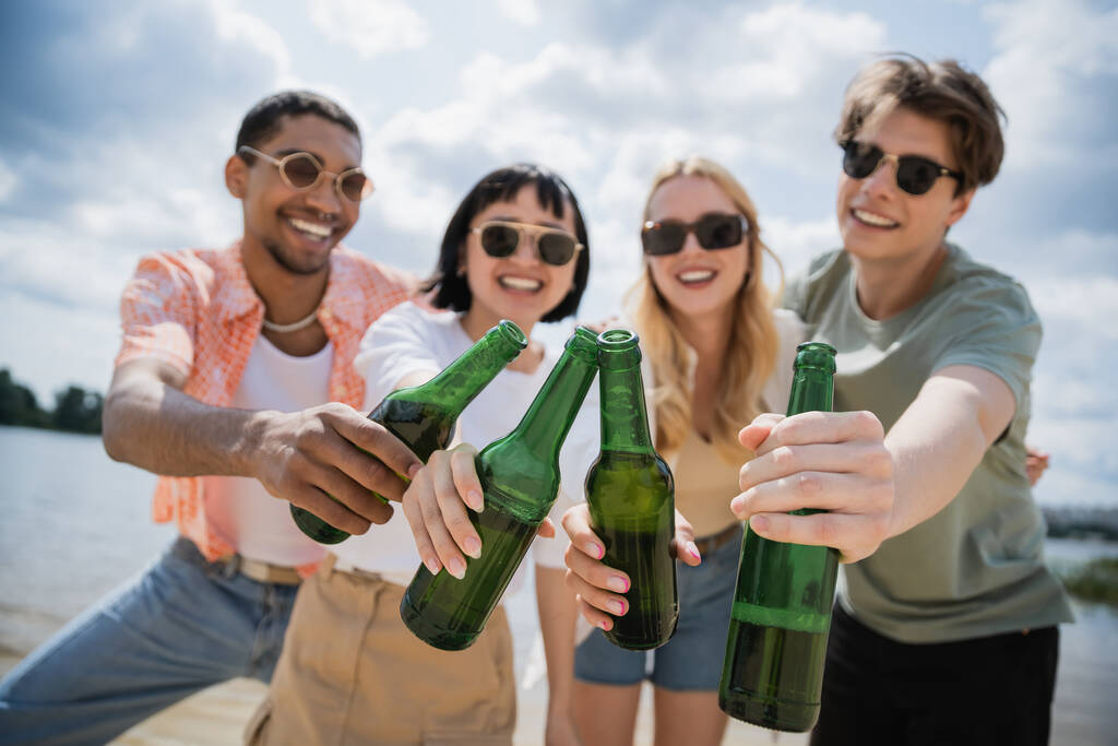 blurred interracial friends in sunglasses clinking bottles during beer party - Photo, image